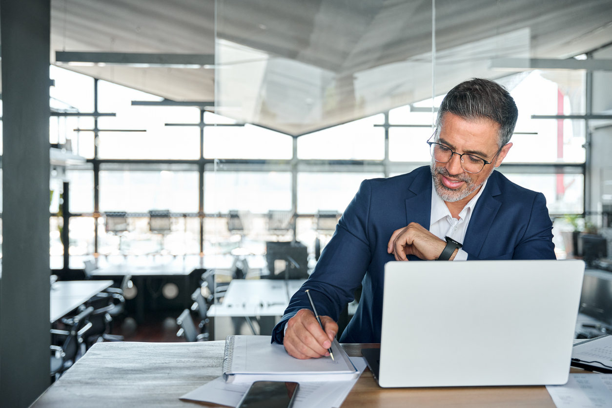 Business man writing notes while on laptop.