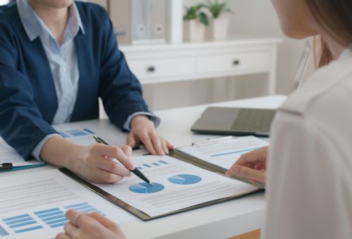Person pointing at chart with pen across the desk from another person.